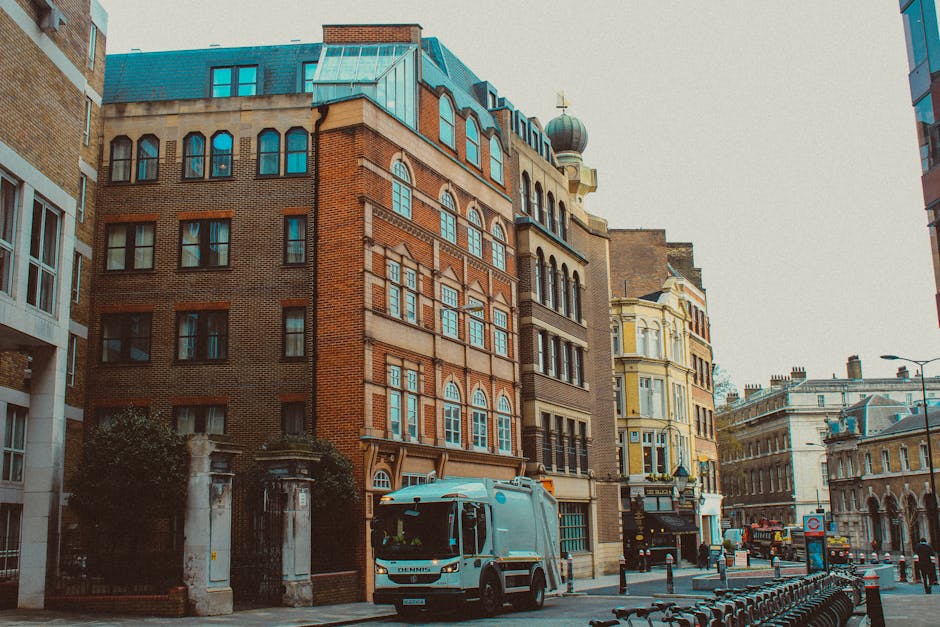 A street scene featuring a white waste collection truck with blue and orange markings parked in front of a row of historic multi-storey buildings, predominantly constructed with red bricks and featuring decorative window frames and arched windows. The buildings display a mix of architectural styles, with some having ornate cornices and rounded bay windows, and the roofs include metal sheeting and small domed structures. The street is partially lined with metal bollards and a bicycle rack in the foreground, with a paved sidewalk alongside. The environment appears to be an urban cityscape during daylight hours, with soft, diffuse lighting indicating overcast weather. Surrounding structures include a yellow-painted building with intricate detailing and a modern glass-and-steel building visible on the right. The scene is indicative of an area where private waste collection and rubbish removal services, such as those provided by Rubbish Clearance Pimlico, might operate, facilitating efficient on-site clearance of waste from urban buildings.