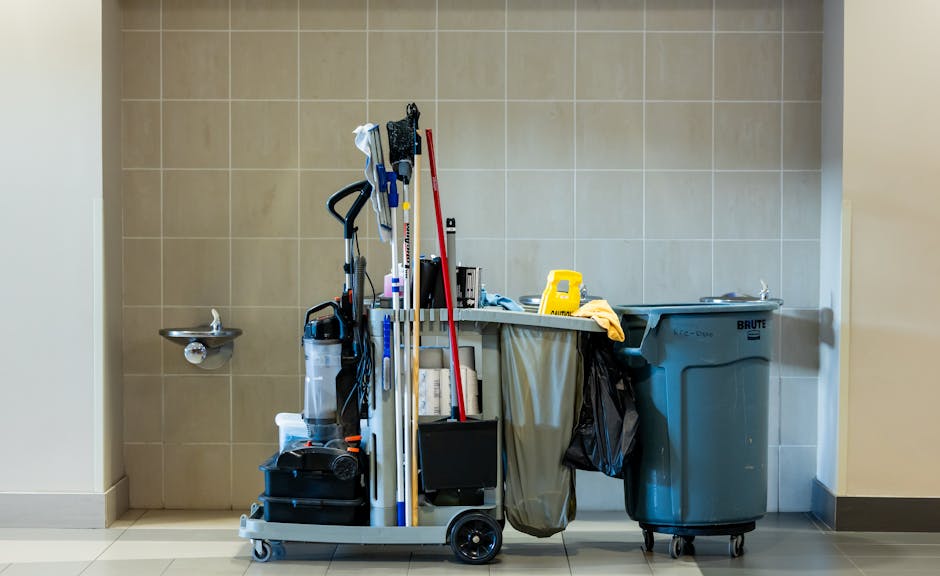 An indoor cleaning and maintenance equipment cart positioned against a beige tiled wall in a commercial or industrial setting. The cart contains various tools and supplies, including a vacuum cleaner, brooms, mops, and cleaning cloths, arranged upright and leaning against each other. A small black box and other cleaning accessories are also visible on the cart's lower shelf. To the right of the cart, there is a large blue waste bin with wheels, labeled as a Brute bin, partially covered with a black plastic bag hanging from its side. Behind the cart, a stainless steel wall-mounted sink with a small faucet is visible, along with a beige wall on the left. The setting appears to be a utility or staff area, possibly within a commercial property, where maintenance tools and waste disposal units are kept for service purposes. The lighting is neutral and evenly distributed, highlighting the practical and functional nature of the space, which may be associated with private waste handling or on-site clearance in a professional environment.