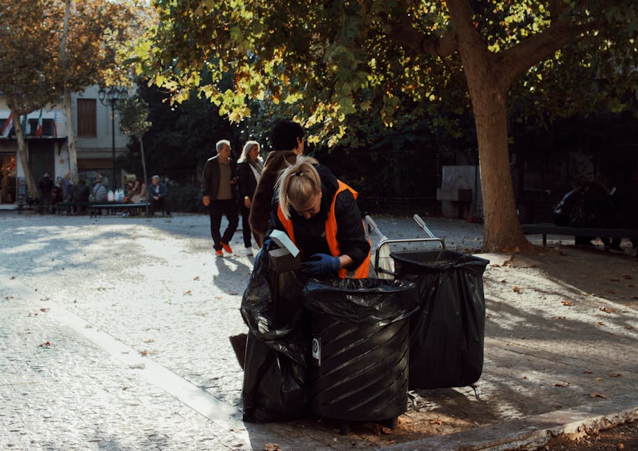 A woman wearing an orange high-visibility vest and blue gloves is bending over to sort waste into large black rubbish bags placed on the cobblestone pavement of a public park or square. The bags are filled with unknown refuse, possibly household or street rubbish, and are positioned next to two wheelie bins, one of which is partially open. In the background, other pedestrians are walking under a canopy of autumn-colored leaves from a large tree, with some people standing near a bench and a building with open storefronts further behind. The scene is illuminated by natural daylight, creating shadows on the ground, and reflects an urban environment where independent waste collection efforts are taking place outside formal municipal services, aligning with private rubbish removal or on-site clearance activities relevant to commercial waste management.