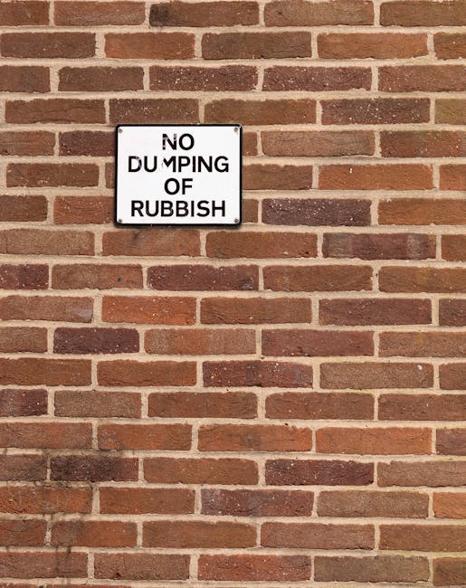 A rectangular metal sign mounted on a red brick wall that reads 'NO DUMPING OF RUBBISH' in black capital letters on a white background. The sign is positioned towards the upper left of the image and is attached with visible screws at the corners. The brick wall features evenly spaced horizontal mortar joints and a consistent pattern of reddish-brown bricks with some darker bricks interspersed, creating a textured surface. Shadows cast by surrounding elements are visible at the bottom left corner of the image, indicating natural daylight. The scene suggests a secure, designated area for waste management, aligning with private rubbish clearance or non-local-authority disposal practices by independent waste services such as Rubbish Clearance Pimlico, which may operate in areas like SW1V or Pimlico.