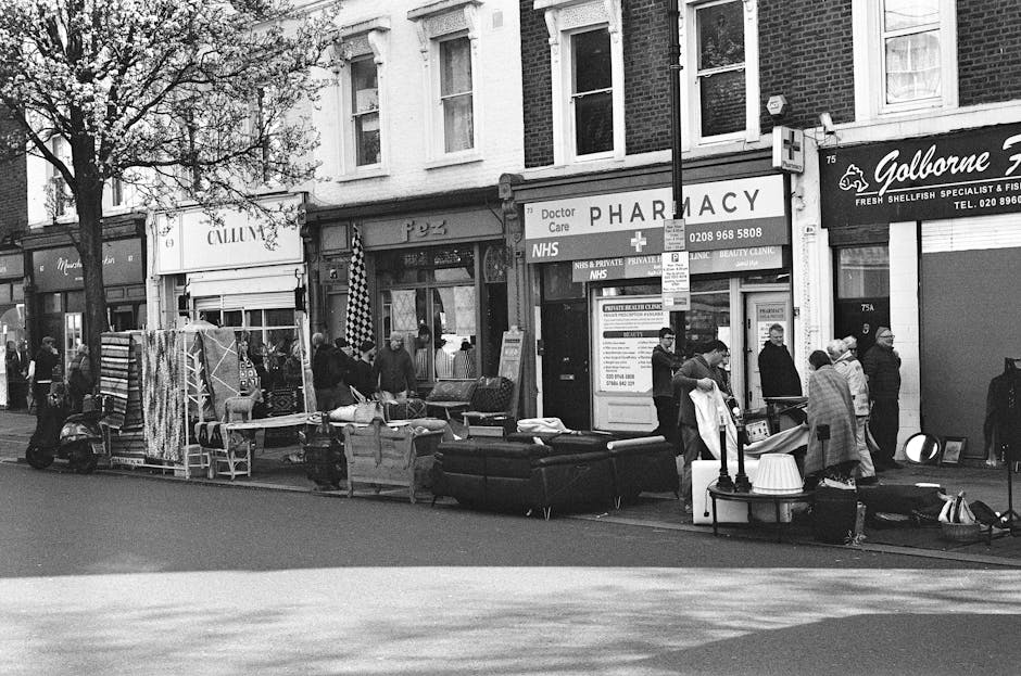 The image depicts a street scene with several people gathered around a variety of discarded furniture and household items placed on the pavement in front of commercial buildings, including a pharmacy and a seafood specialist shop. Visible objects include a dark-colored sofa, a mattress, a small table lamp, and some smaller household goods, all appearing to be used and casually arranged. The furniture displays textures typical of fabric and upholstered surfaces, with the sofa and mattress showing indications of wear. The surrounding environment features a row of brick-fronted shops, with signage for a pharmacy and other retail outlets, and a large tree on the left side partially shading the pavement. The scene is lit by natural daylight, with subtle shadows cast by the gathered individuals and items. This outdoor scene reflects a situation involving private waste collection or informal rubbish disposal, consistent with services provided by rubbish clearance specialists such as Rubbish Clearance Pimlico, offering alternatives to official waste disposal methods.