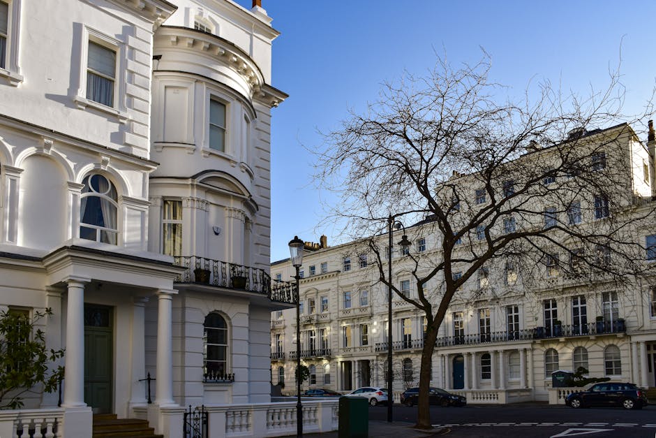 A street scene featuring a white, Victorian-style residential building with decorative architectural details, including columns, bay windows, and a small balcony with black iron railings. The building's façade is smooth with a glossy finish, contrasting with the dark window frames. To the right, there is a leafless tree with thin, twisting branches that extend across the sky, which is clear and blue. In the background, there are additional similar residential buildings with uniform white facades, multiple windows, and black balconies, creating a consistent architectural style typical of urban Victorian terraces. A row of parked cars lines the street, and a lamppost stands near the tree, adding to the street’s urban character. The scene is well-lit, with natural daylight highlighting the details of the buildings and tree, offering a clear view suitable for illustrating private waste collection or property clearance contexts in an urban environment. Rubbish Clearance Pimlico services could relate to maintaining such areas through efficient rubbish removal for residential properties.