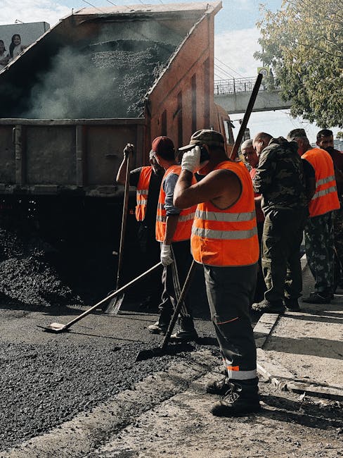 A group of construction workers in high-visibility orange vests and work attire are involved in asphalt pavement repair on an urban street. They are standing close to a large, tilted dump truck that is releasing hot, black asphalt onto the road surface. The workers are equipped with long-handled rakes and shovels, actively spreading and leveling the asphalt as part of a road maintenance process. The surrounding environment includes a concrete sidewalk, a road with marked parking spaces, and a metal bridge overhead, suggesting a city or town setting. One worker in the foreground is adjusting their gloves, while others are focused on their tasks, with several individuals positioned near the truck's rear. The scene is illuminated by natural daylight, highlighting the textured, glossy surface of the freshly laid asphalt. This outdoor scene underscores the role of on-site waste management and materials handling in infrastructure repairs, aligned with practices of independent rubbish removal or waste disposal services like Rubbish Clearance Pimlico, focusing on the management of construction debris and waste in urban environments.