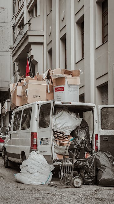A white vans with open rear doors parked on a city street in front of a multi-storey building with beige stone facade and large windows. The van's interior is filled with black and white plastic rubbish bags, cardboard boxes, and other miscellaneous waste materials, some of which are piled and leaning against the vehicle's sides. Several large cardboard boxes, some open and partially collapsed, are stacked on top of the van, with others resting on the pavement nearby. The scene includes a small hand trolley loaded with black refuse bags and a few loose waste items in the foreground. The environment appears to be an urban area with a narrow street and minimal street furniture visible. This setup illustrates independent waste collection or private rubbish clearance, which could be part of a professional rubbish removal service such as Rubbish Clearance Pimlico, serving residential or commercial premises that require on-site clearance outside conventional council collection. The overall scene emphasizes the practical aspects of on-demand rubbish disposal in a city environment, with a focus on waste containment and transportation readiness.