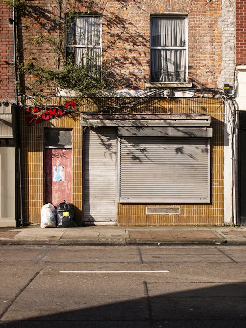 A ground-floor storefront in an urban setting featuring a closed, grey metal roll-up shutter with visible horizontal ridges, set within a building façade clad in brown and yellow tiles. To the left of the shutter, there are two black garbage bags with some white plastic bags placed next to them, and a red and pink boarded-up window or door with graffiti markings beneath the left window. Above, the building's upper story includes two centrally aligned sash windows with white frames, with curtains partially drawn, and brickwork with a weathered, reddish-brown appearance. Green foliage from a nearby tree casts shadows onto the brick wall, adding contrast to the scene. The pavement in front shows some staining and minor surface cracks, typical of city streets. This scene subtly reflects private waste or rubbish removal practices often associated with independent collection or site clearance, aligning with the services offered by Rubbish Clearance Pimlico in managing rubbish disposal in the area.