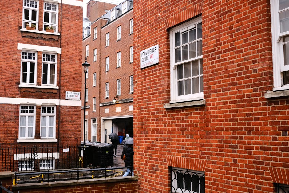 A white vans with open rear doors parked on a city street in front of a multi-storey building with beige stone facade and large windows. The van's interior is filled with black and white plastic rubbish bags, cardboard boxes, and other miscellaneous waste materials, some of which are piled and leaning against the vehicle's sides. Several large cardboard boxes, some open and partially collapsed, are stacked on top of the van, with others resting on the pavement nearby. The scene includes a small hand trolley loaded with black refuse bags and a few loose waste items in the foreground. The environment appears to be an urban area with a narrow street and minimal street furniture visible. This setup illustrates independent waste collection or private rubbish clearance, which could be part of a professional rubbish removal service such as Rubbish Clearance Pimlico, serving residential or commercial premises that require on-site clearance outside conventional council collection. The overall scene emphasizes the practical aspects of on-demand rubbish disposal in a city environment, with a focus on waste containment and transportation readiness.