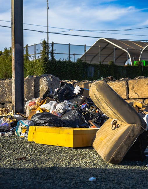 A heap of mixed household and construction waste is piled on the pavement beside a low stone wall. The debris includes black garbage bags, loose plastic packaging, and discarded items such as an old, dusty beige car seat or cushion with visible fabric wear and slight tears. A bright yellow plastic container or tray is partly underneath the bags, with other smaller plastic items scattered around. In the background, there is a metal pole with overhead wires running across the sky, which is partly cloudy with patches of blue. Behind the waste, a dark green hedge provides a boundary, and further behind, there is a large outdoor sports or tennis court covered by a white tensile fabric roof structure. The scene appears to be taken during daylight hours with natural lighting highlighting the textures of the waste, emphasizing the importance of professional rubbish removal services like those offered by Rubbish Clearance Pimlico for private disposal or on-site clearance needs.