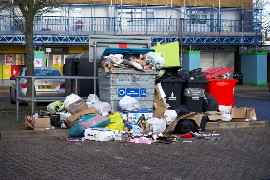 A collection of overflowing rubbish bins and scattered waste bags on a paved pavement outside a commercial building. The waste includes cardboard boxes, plastic bags, paper, and packaging materials, some of which are torn or crumpled. The grey and black bins are partially open, revealing various discarded items, while a bright red bin stands nearby. Surrounding the waste are small cardboard boxes and loose paper strewn across the ground. In the background, there is a modern building with a scaffolding-covered facade and a row of shopfronts, some with signs indicating food services. A tree with bare branches is positioned to the left of the rubbish, and a parked silver car is on the pavement behind a metal railing. Natural daylight illuminates the scene, highlighting the disorder and suggesting a need for waste collection or rubbish removal services, such as those offered by Rubbish Clearance Pimlico.