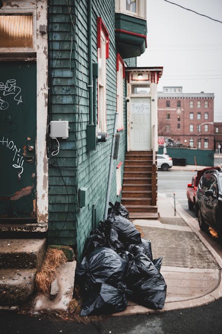 A collection of several black rubbish bags piled against the green wooden exterior wall of a building on a city street. The bags are tightly tied and stacked on a concrete sidewalk next to a small patch of ice and a patch of dried grass near a worn concrete staircase leading up to a white door with a red and white striped awning. The building has an exterior of weathered green wooden shingles, with utility boxes and cables attached to the wall. In the background, a parked black car is visible along with other vehicles on a paved street, and a large brick building can be seen further in the distance under an overcast sky. The scene appears to depict independent rubbish collection possibly arranged by a private waste management service, with the rubbish awaiting removal from the premises, aligning with alternative waste handling approaches.
