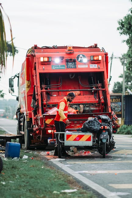 A sanitation worker, wearing a high-visibility orange jacket and black trousers, is operating a large red rubbish collection vehicle on a city street. The rear loading mechanism of the waste truck is open, revealing the internal compactor, which appears to be in active use. The worker is standing beside a black trash bag placed on a metal platform attached to the vehicle, and there are scattered bits of waste around the area. The scene occurs during daytime, with an overcast sky, and the environment includes a curbside with a strip of grass, a bicycle parked nearby, and some low-rise buildings visible in the background. Utility poles and power lines run along the street, contributing to the urban setting. The scene illustrates a routine rubbish collection operation, reflecting private waste handling methods that supplement or serve as alternatives to municipal services, aligning with the concept of independent waste disposal and rubbish removal in Pimlico.