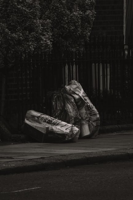 A narrow urban alleyway cluttered with various types of discarded rubbish, including a large black and white bag in the foreground, which appears to be filled with waste and is placed on a rough, uneven concrete surface. Behind the bag, there are multiple stacked cardboard boxes, plastic bags, and miscellaneous debris piled haphazardly, filling the alley to the point of overflowing onto the surrounding area. The alley is flanked on both sides by brick and metal buildings, with graffiti visible on the walls to the right. Bare tree branches extend overhead, and the scene appears dimly lit, suggesting overcast weather or late afternoon lighting. A small black waste bin is positioned near the left side of the alley, and a trolley or cart with a metal handle is partly visible behind the large bag. This scene exemplifies the type of setting where private rubbish clearance services, such as those offered by Rubbish Clearance Pimlico, might operate to aid in cleaning and waste removal from urban environments, though no equipment or collection vehicle is present in the image itself.